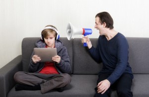 Tween son in headphones looks at the digital tablet display while his father yells at him through a megaphone